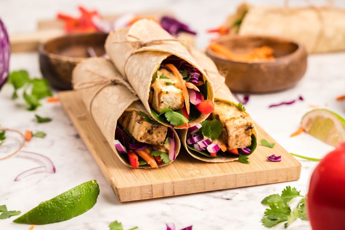 Three vegan and gluten free wraps, filled with tofu and veggies, are tied with twine on a wooden board, surrounded by fresh herbs and lime wedges—perfect as an easy tofu wrap to pair with Coconut Almond Butter Dip.