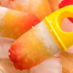 Colorful Coconut ice pops with plastic handles rest on ice cubes in a wooden tray, perfect for a refreshing treat.