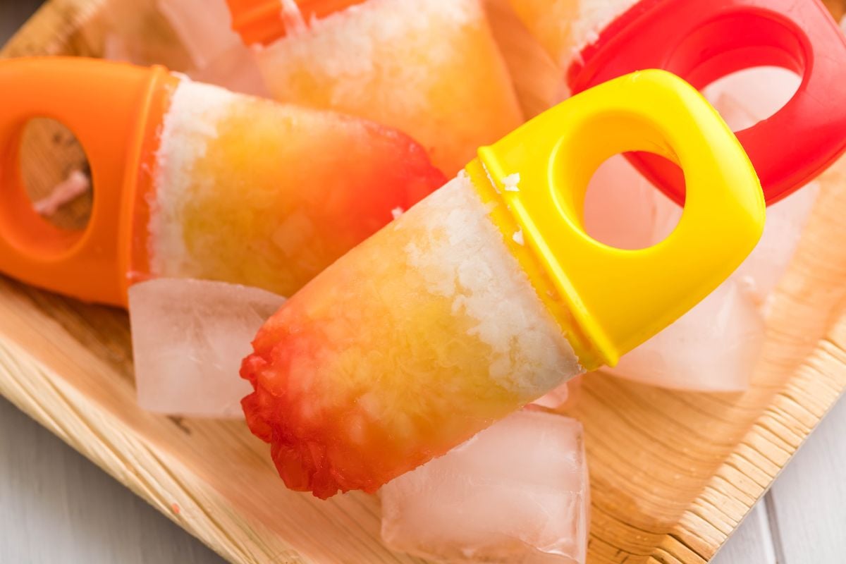Colorful homemade sugar free popsicles with ice cubes on a wooden plate, featuring orange and yellow handles—perfect for trying your own layered popsicle recipe.