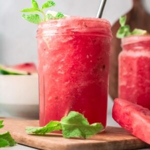 Two mason jars filled with a frothy pink Watermelon White Wine Slushie, garnished with mint, sit beside watermelon slices—a refreshing frozen watermelon cocktail perfect for summer.