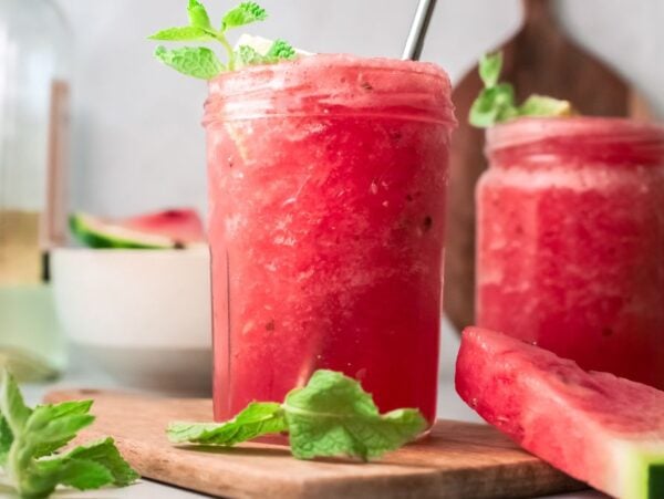 Two mason jars filled with a frothy pink Watermelon White Wine Slushie, garnished with mint, sit beside watermelon slices—a refreshing frozen watermelon cocktail perfect for summer.