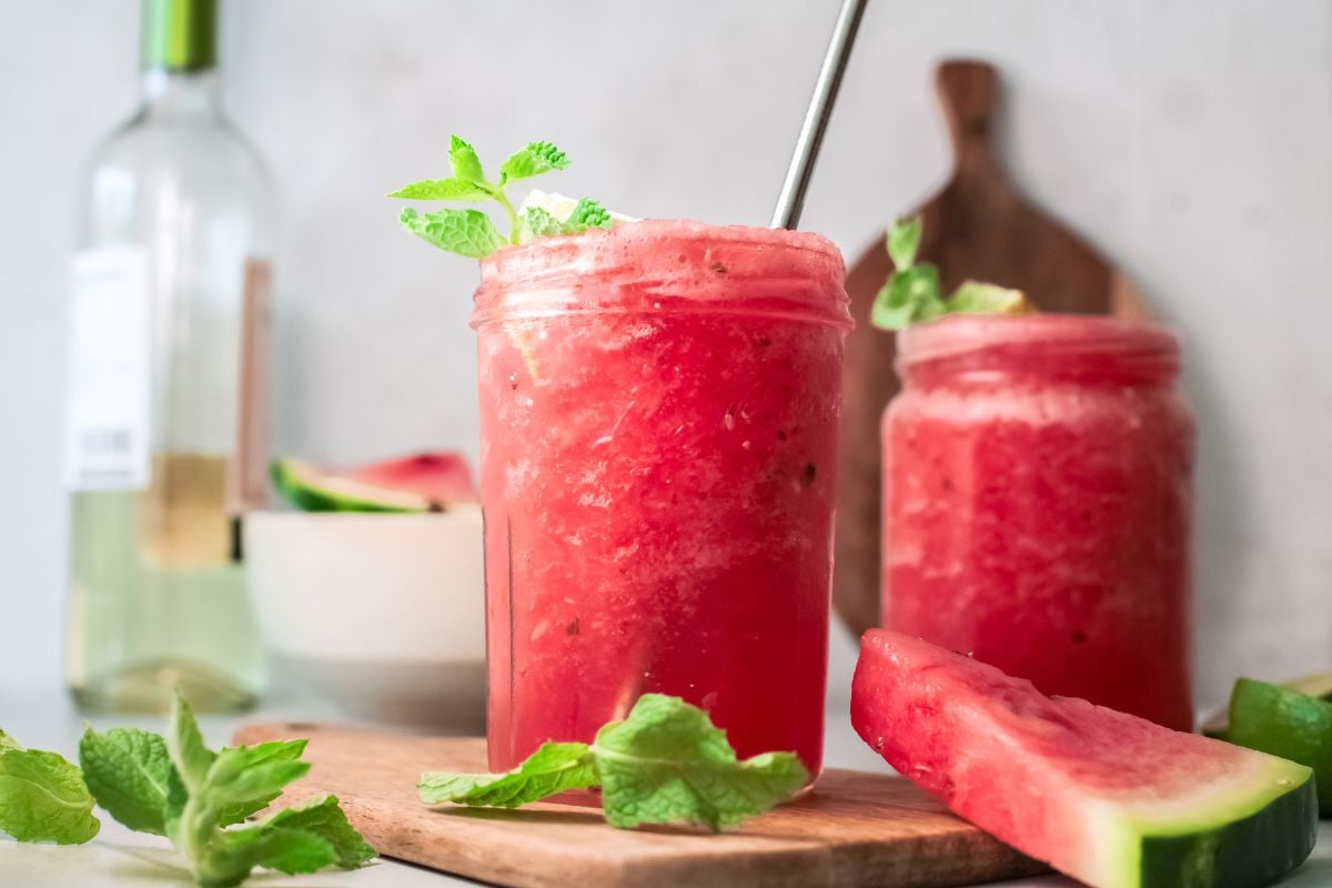 Two mason jars of Frozen Watermelon Cocktail with mint, a metal straw, and watermelon slices on a wooden board.