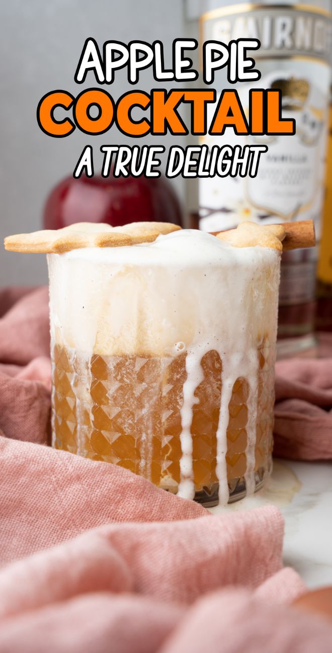 A frothy apple pie cocktail made with apple cider in a glass, topped with pie crust, with bottles and a red apple in the background. This autumn cocktail recipe is perfect for cozy fall gatherings.
