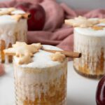 Three glasses of frothy Apple Pie Cocktail, made with apple cider and topped with cinnamon sticks and star-shaped cookies, are surrounded by fresh apples.