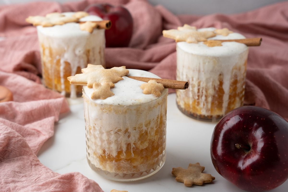 Three glasses of frothy apple cinnamon cocktail, topped with cinnamon sticks and leaf cookies, with apples in the background.