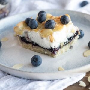 A Blueberry Pie Meringue Square topped with blueberries and almond slices sits on a light blue plate, with a fork in the background—an irresistible bar recipe with meringue for any easy blueberry dessert craving.