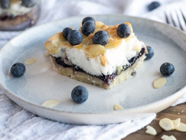 A Blueberry Pie Meringue Square topped with blueberries and almond slices sits on a light blue plate, with a fork in the background—an irresistible bar recipe with meringue for any easy blueberry dessert craving.