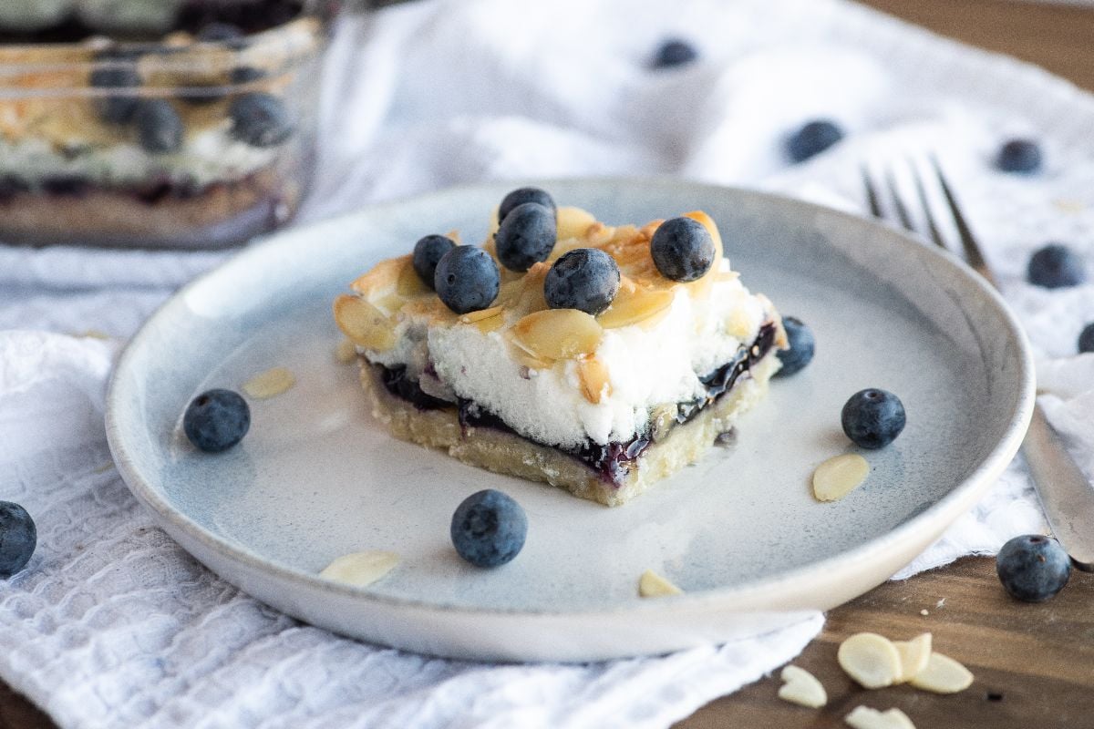 A dessert bar topped with blueberries and almond slices on a plate, featuring almond and blueberry cookie bars, with more blueberries scattered around.