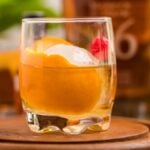 A classic bourbon cocktail, this drink features an orange peel, cherry, and ice ball in a glass on a wooden tray—a timeless bourbon Old Fashioned you can make at home.