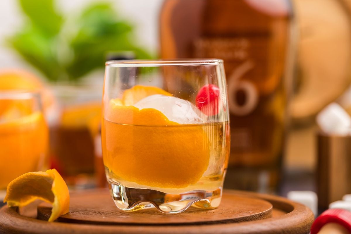 A glass of whiskey with ice, an orange peel, and a cherry on a wooden tray, with a whiskey bottle in the background—showcasing how to make a bourbon old fashioned and master the best old fashioned recipe.