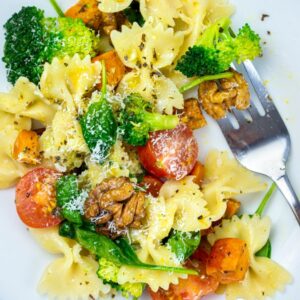 A plate of vegetable pasta salad with bowtie pasta, broccoli, cherry tomatoes, walnuts, and grated cheese, served with a fork.