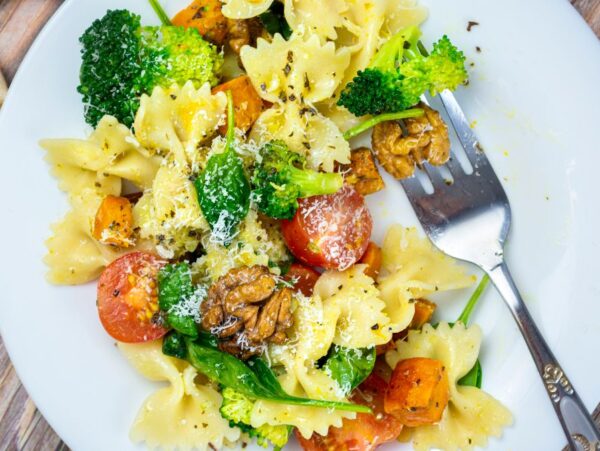 A plate of vegetable pasta salad with bowtie pasta, broccoli, cherry tomatoes, walnuts, and grated cheese, served with a fork.