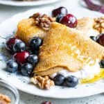 A sweet and savoury crepe with cream, blueberries, cherries, walnuts, and honey on a white plate with floral patterns—perfect for those who crave flavourful crepes for brunch.