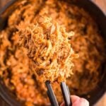 Tongs holding shredded pulled pork in a Dutch oven over a pot filled with more pulled pork, perfect to pair with Dairy Free Vegan Coleslaw on a wooden table.