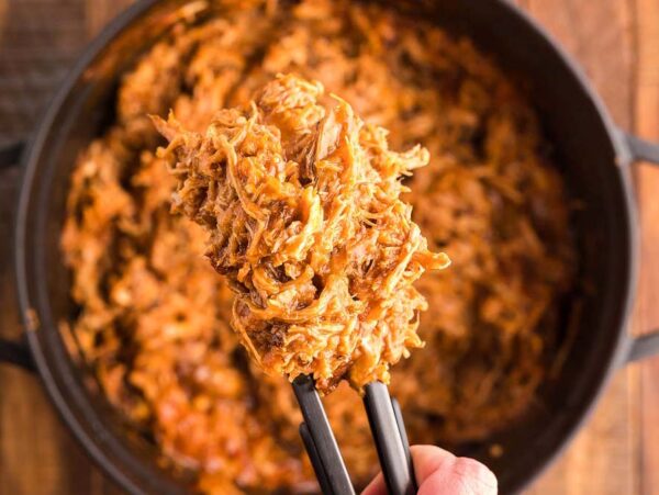 Tongs holding shredded pulled pork in a Dutch oven over a pot filled with more pulled pork, perfect to pair with Dairy Free Vegan Coleslaw on a wooden table.