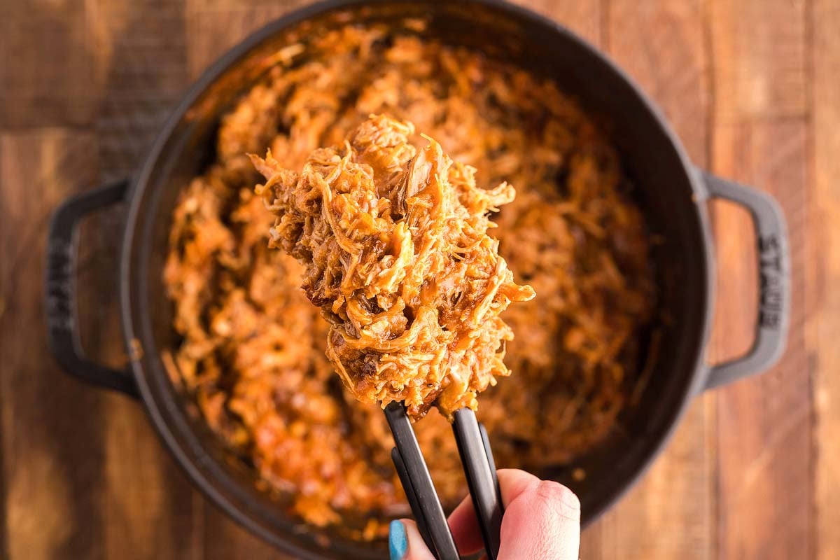 Hand holding tongs with shredded BBQ pulled pork in a dutch oven over a pot on a wooden table.