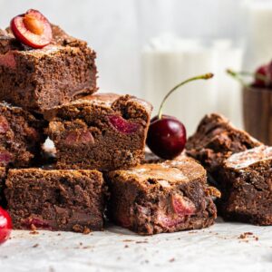 A stack of Fresh cherry brownies with juicy cherries, surrounded by fresh cherries and glasses of milk in the background—an easy cherry dessert perfect for any occasion.