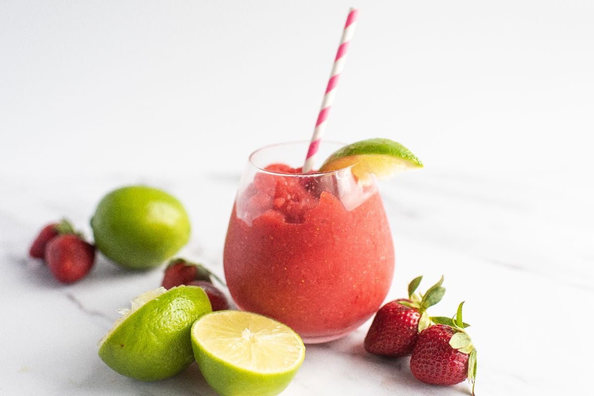 A glass of frozen strawberry lime daiquiri with a striped straw, garnished with lime, surrounded by strawberries and limes—an easy way to make a daiquiri everyone will love.