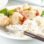 Plate of white rice with grilled pepper and chicken kebabs, served alongside vegetables, garnished with parsley, and a fork on the side.