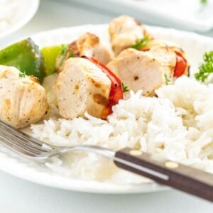 Plate of white rice with grilled pepper and chicken kebabs, served alongside vegetables, garnished with parsley, and a fork on the side.
