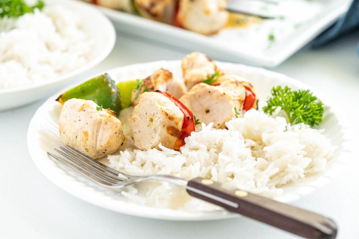 Plate of white rice and grilled pepper and chicken kebabs with vegetables, garnished with parsley, and a fork on the side.