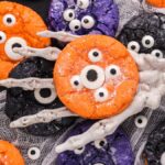A skeleton hand holds an orange monster cookie with candy eyes; Halloween Crinkle Cookies and other colorful cookies are nearby.