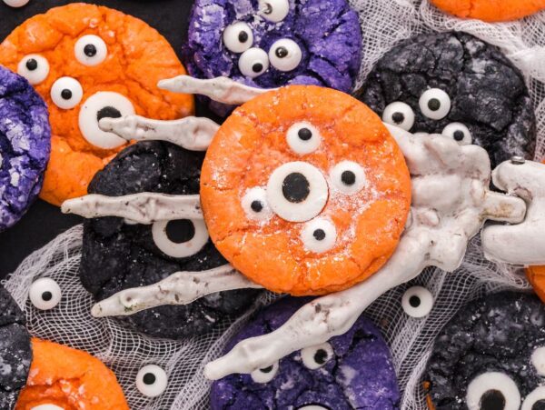 A skeleton hand holds an orange monster cookie with candy eyes; Halloween Crinkle Cookies and other colorful cookies are nearby.