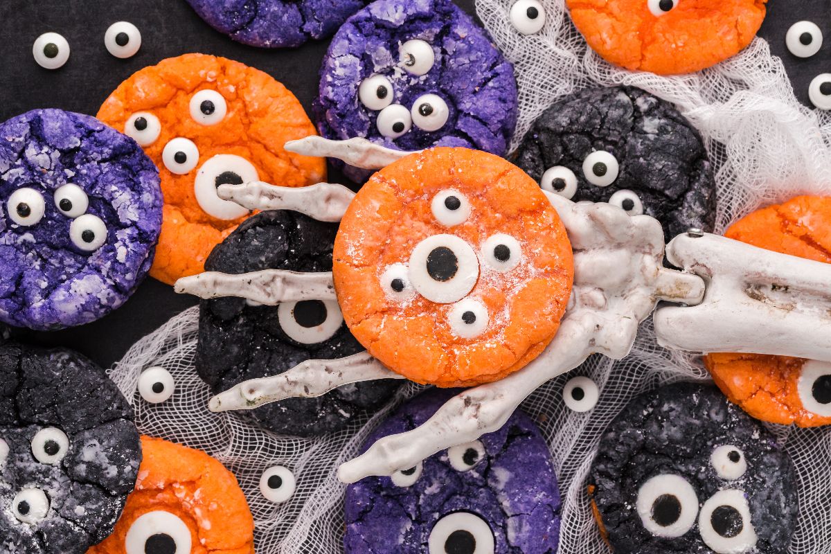 Colourful monster cookies with candy eyes, held by a skeleton hand, set on a festive mesh background&mdash;perfect inspiration for easy Halloween cookie decorating ideas.