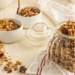 A glass jar and bowls filled with granola, nuts, and oats on a light table with a striped towel—perfect for anyone seeking easy homemade granola as part of a healthy breakfast.