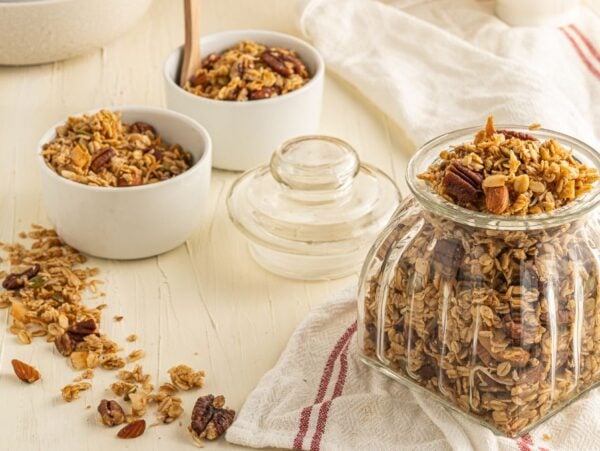 A glass jar and bowls filled with granola, nuts, and oats on a light table with a striped towel—perfect for anyone seeking easy homemade granola as part of a healthy breakfast.