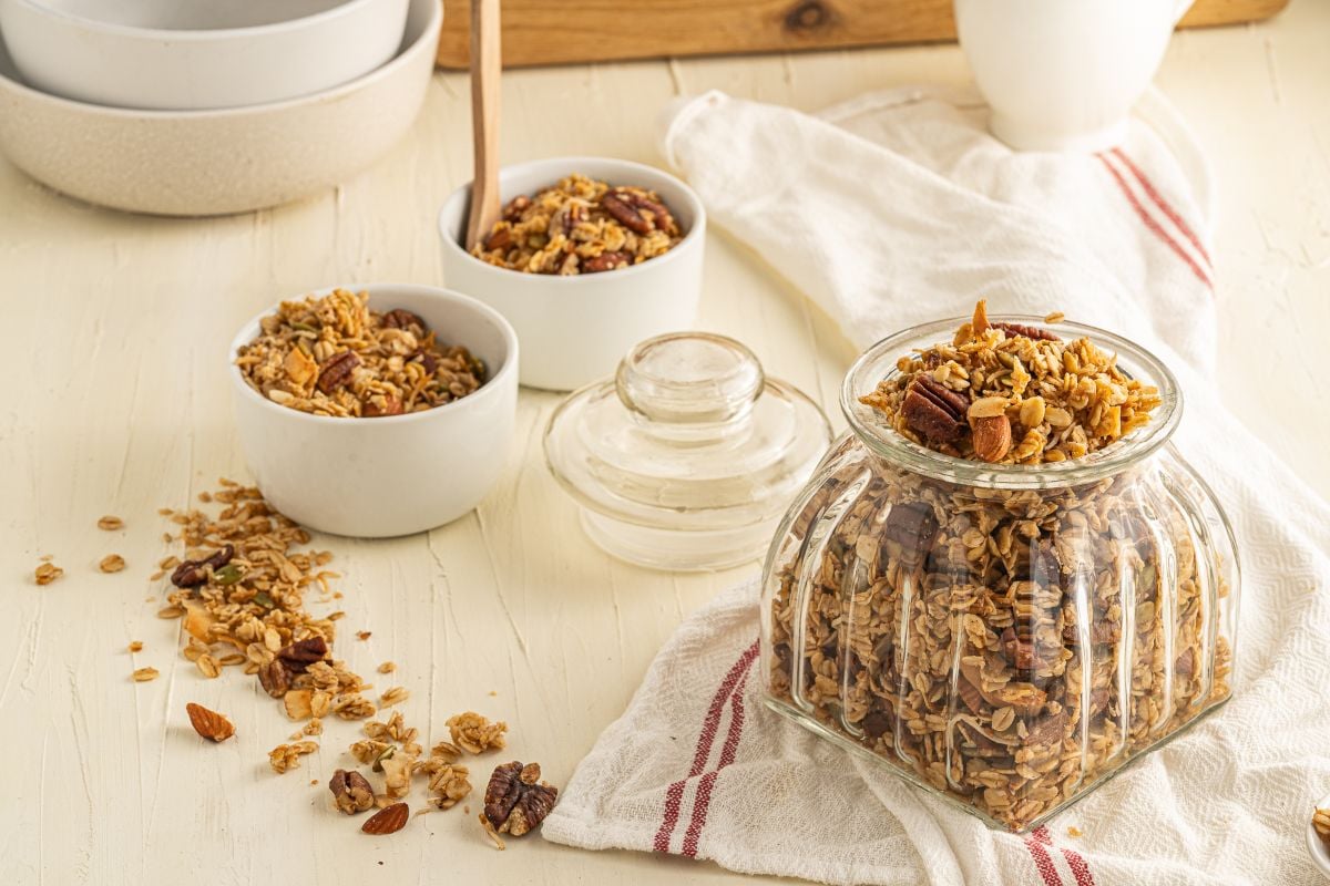 Jar and bowls of easy homemade granola on a table with spilled granola and a white cloth nearby—perfect for anyone looking to learn how to make granola at home.