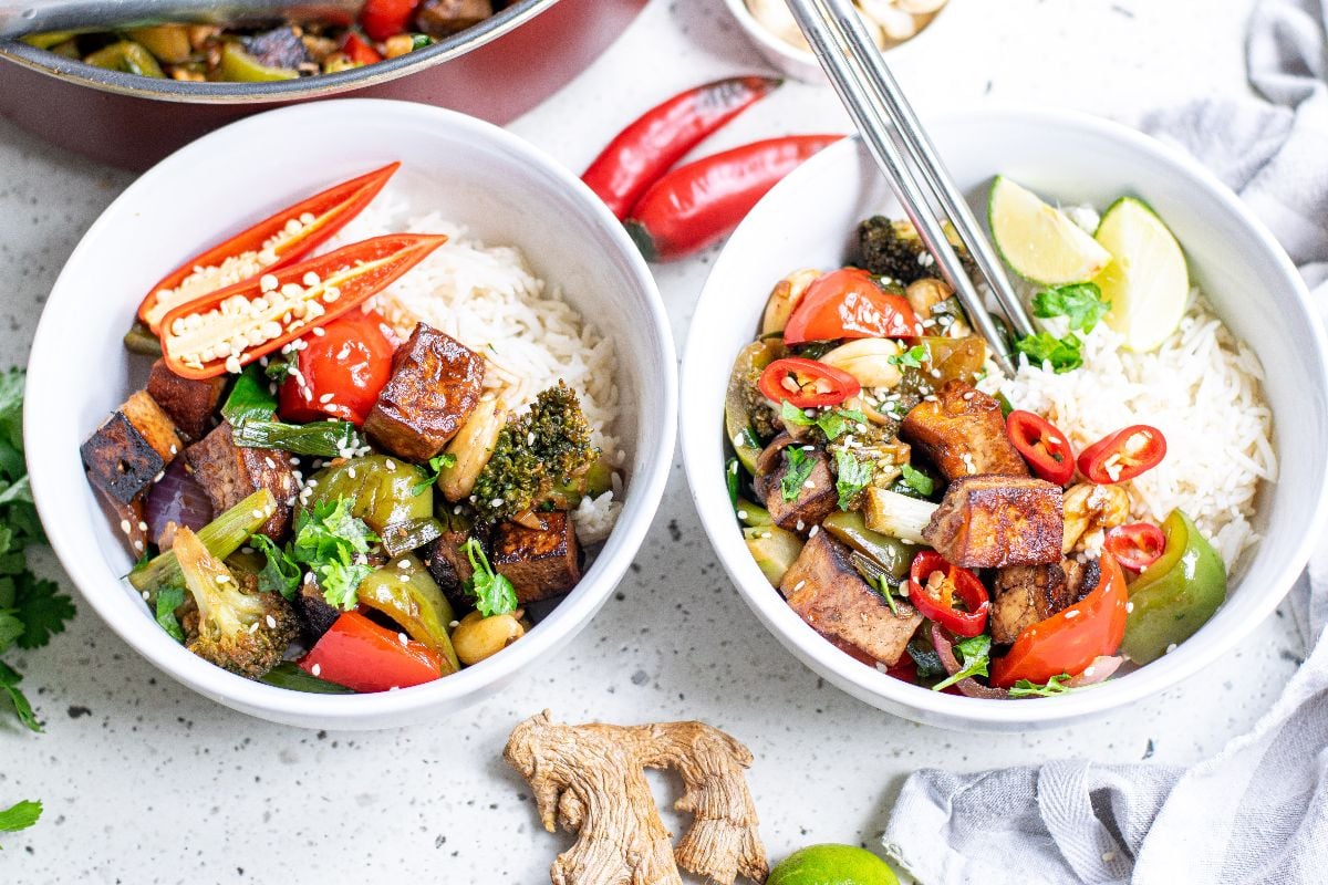 Two bowls of rice topped with Kung Pao Tofu, stir-fried vegetables, red chili slices, and lime wedges for a delicious Vegetarian Chinese inspired dinner.