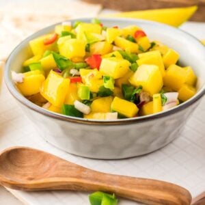 A bowl of vibrant mango salsa with diced mango, peppers, onions, and herbs sits next to flatbread and a red chili pepper—a perfect summer salsa recipe for any gathering.