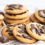 A stack of chocolate-drizzled cookies with chunks of chocolate sits on a light cloth surface—perfect inspiration for anyone wanting to learn how to make stuffed cookies or try tasty Mars bar stuffed cookies at home.