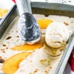 Vanilla ice cream with peach slices in a pan, an ice cream scoop, and a bowl of peaches nearby—this no churn peach ice cream is perfect for summer treats.