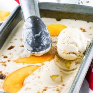 Vanilla ice cream with peach slices in a pan, an ice cream scoop, and a bowl of peaches nearby—this no churn peach ice cream is perfect for summer treats.