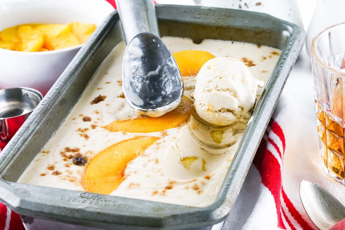 A metal pan with No Churn Bourbon Peach Ice Cream being scooped, topped with peach slices and brown crumbs.