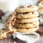 A stack of chocolate chip cookies, one broken open, with milk and peanut butter cups in the background—these are irresistible peanut butter cup stuffed cookies.