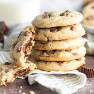 A stack of chocolate chip cookies, one broken open, with milk and peanut butter cups in the background—these are irresistible peanut butter cup stuffed cookies.