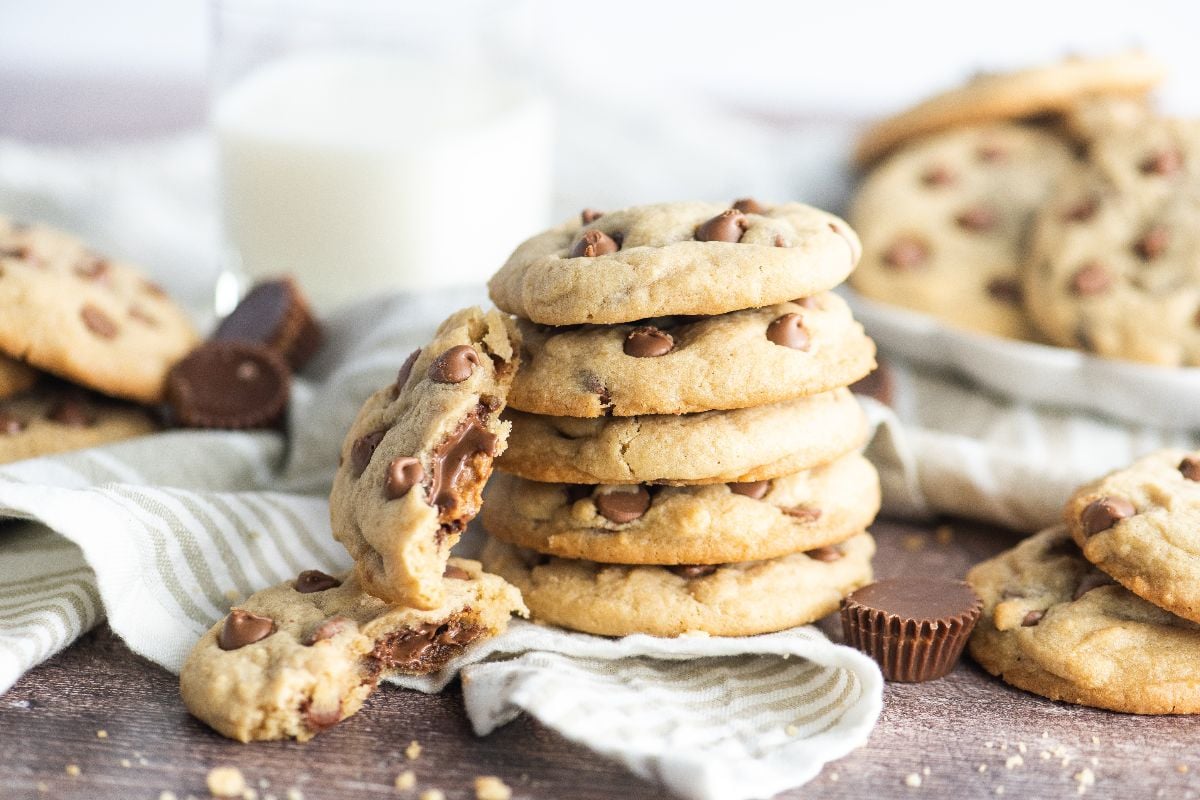 A stack of peanut butter cup stuffed cookies, with one broken in half, sits next to a glass of milk—each cookie made with mini peanut butter cups.
