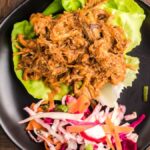 Pulled pork in lettuce wraps with vibrant dairy free coleslaw sits on a black plate atop a wooden table, showcasing tender slow cooked pork shoulder and fresh flavors.