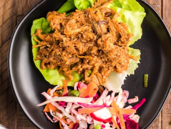 Pulled pork in lettuce wraps with vibrant dairy free coleslaw sits on a black plate atop a wooden table, showcasing tender slow cooked pork shoulder and fresh flavors.