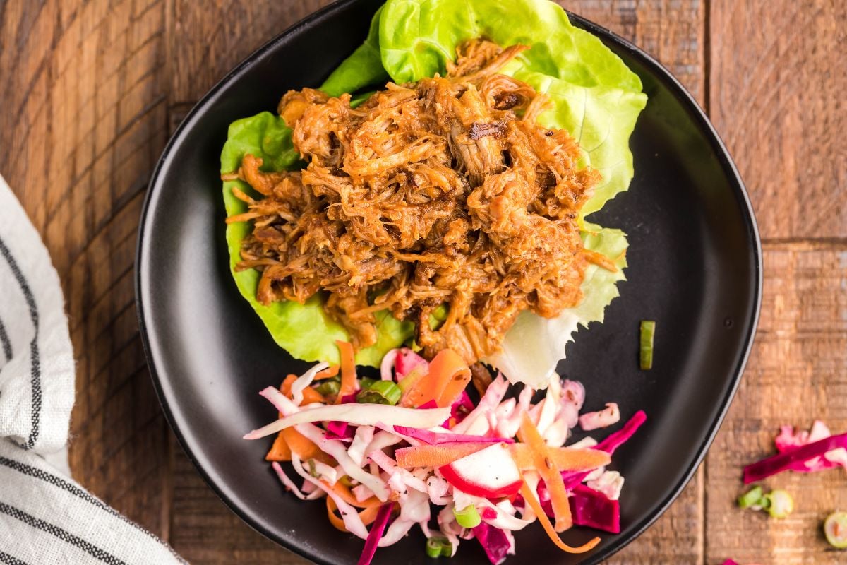 Pulled pork in lettuce wraps with vibrant, dairy free coleslaw on a black plate, set atop a wooden table.