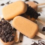 Four caramel pumpkin popsicles, two topped with an Oreo coating, on a white surface with cookie crumbs—a delicious twist for any pumpkin spice lover.