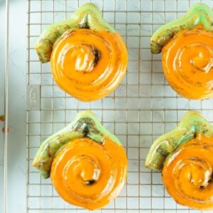Six Pumpkin Shaped Cinnamon Rolls with orange icing cool on a rack, next to an apple in a wooden bowl—a festive pumpkin shaped dessert idea made from Pillsbury Grand Cinnamon Rolls.