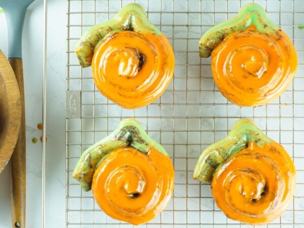 Six Pumpkin Shaped Cinnamon Rolls with orange icing cool on a rack, next to an apple in a wooden bowl—a festive pumpkin shaped dessert idea made from Pillsbury Grand Cinnamon Rolls.