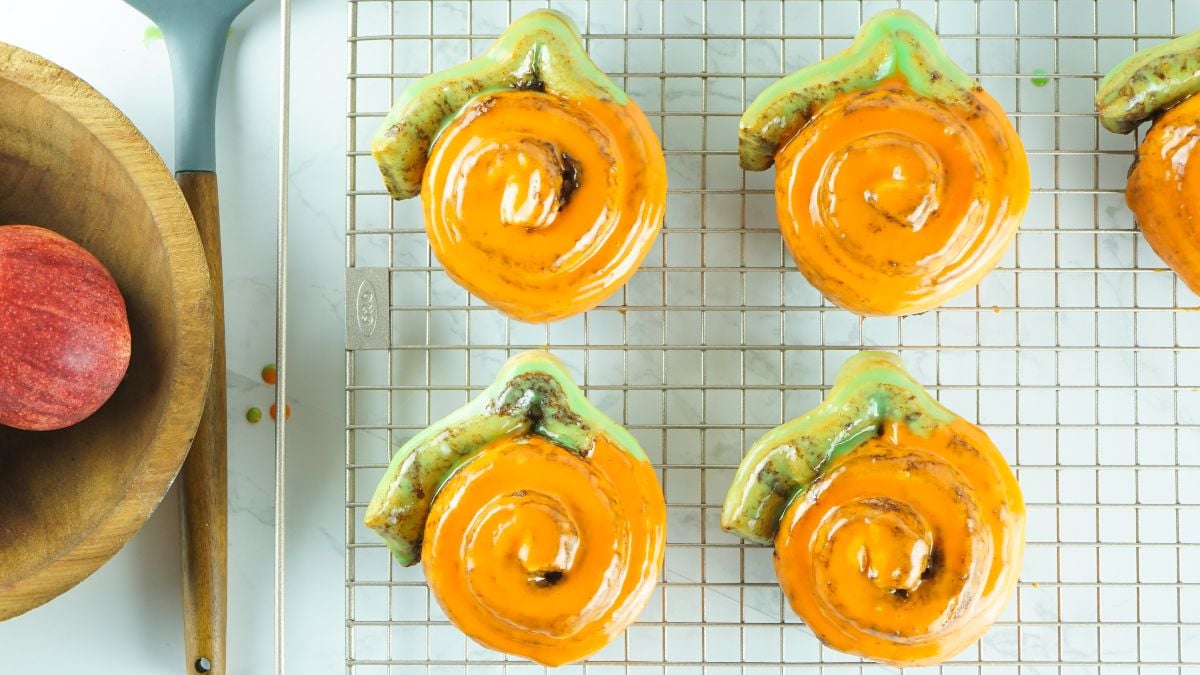 Six Pumpkin Shaped Cinnamon Rolls with orange icing cool on a rack, next to an apple in a wooden bowl—a festive pumpkin shaped dessert idea made from Pillsbury Grand Cinnamon Rolls.