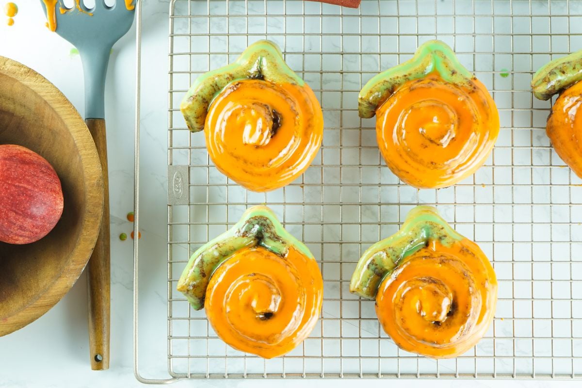 Six Pumpkin Shaped Cinnamon Rolls made from Pillsbury Grand Cinnamon Rolls, with orange icing, rest on a cooling rack beside a spatula and an apple—perfect homemade pumpkin treats for fall.