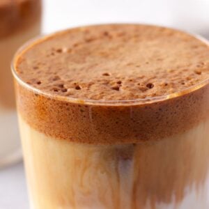 Glass of dalgona coffee with pumpkin whipped coffee foam on top and milk underneath, made with pumpkin puree and instant coffee, evoking an autumn coffee flavour. Blurred background.