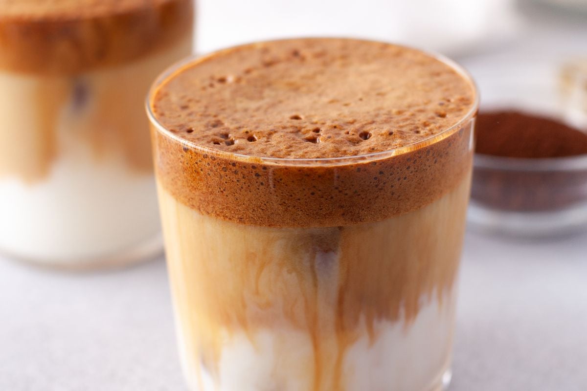 A glass of Pumpkin Spice Whipped Dalgona Coffee with creamy foam on top and milk beneath, perfect for a cozy seasonal coffee recipe, sits on a light surface.